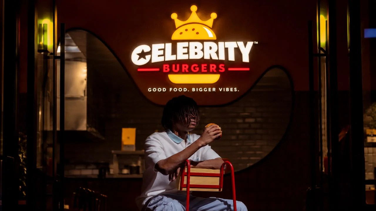 A lit Celebrity Burgers sign above a person eating a burger on a red chair