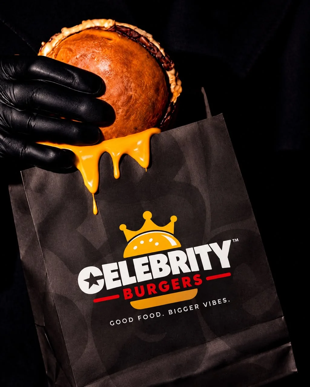 A gloved hand holding a burger over a black Celebrity Burgers paper bag, with cheese dripping onto it